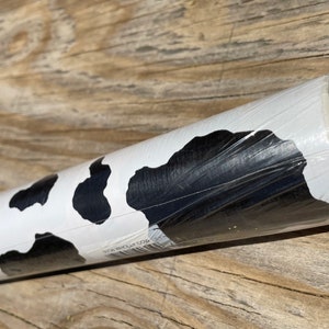 May include: A roll of black and white cow print contact paper. The paper is wrapped around a cardboard tube and is sealed in plastic.