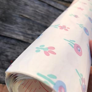 May include: Rolled-up white paper with a repeating floral pattern in pink, blue, and teal. The paper has red measurement markings. The paper is rolled up, revealing the inside layers.