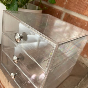 May include: A clear plastic drawer organizer with three drawers and silver knobs. The organizer is empty.