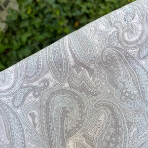 May include: Close-up of a fabric with a paisley pattern. The fabric features a repeating design of gray and light blue paisley shapes on a white background. The fabric is folded, revealing the pattern's intricate details.