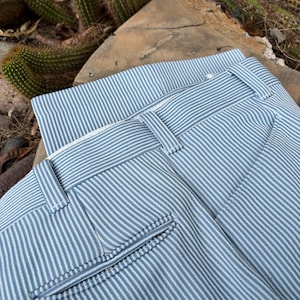 May include: Close-up of blue and white striped pants. The pants have a classic design with a button and zipper closure, belt loops, and a pocket. The fabric has a vertical stripe pattern.
