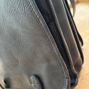 Puede incluir: Primer plano de un bolso de cuero negro. El bolso tiene una superficie texturizada y un cierre de solapa con hebilla. Las costuras son visibles a lo largo de los bordes. El bolso parece desgastado y tiene un dise&ntilde;o cl&aacute;sico.