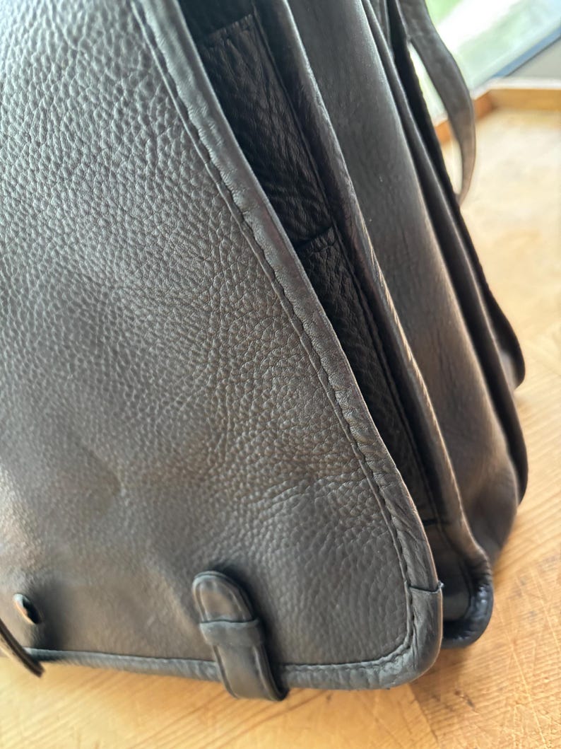 Puede incluir: Primer plano de un bolso de cuero negro. El bolso tiene una superficie texturizada y un cierre de solapa con hebilla. Las costuras son visibles a lo largo de los bordes. El bolso parece desgastado y tiene un dise&ntilde;o cl&aacute;sico.