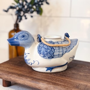 Peut inclure: Une théière en céramique en forme de canard avec une base blanche et des détails peints à la main en bleu sur la tête et les ailes. La théière dispose d'une anse en rotin tressé marron clair fixée à deux petites boucles sur le dessus.