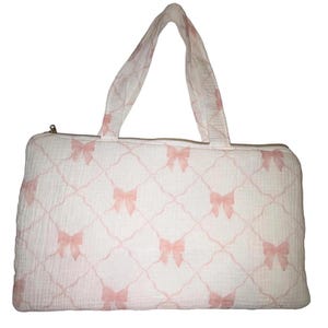 May include: A rectangular tote bag made of white fabric with a soft pink lattice pattern and small pink bows at each intersection. The bag features two top handles and a zipper closure along the top edge, showcasing a textured, lightweight material.
