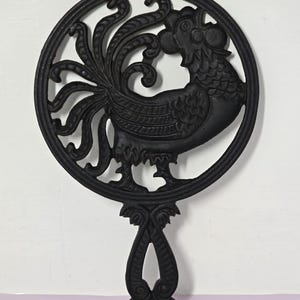 Puede incluir: Un salvamanteles circular de hierro fundido negro con un diseño calado de un gallo con cola y cresta detalladas. El salvamanteles incluye un mango decorativo que se extiende desde la parte inferior del marco circular con volutas.
