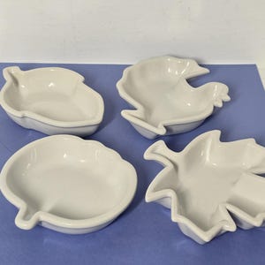 Może przedstawiać: Zestaw czterech białych ceramicznych miseczek o różnych kształtach, w tym żołędzia, kurczaka, dyni i liścia klonu. Każde naczynie ma gładkie, błyszczące wykończenie z podniesionymi krawędziami, przeznaczone do serwowania niewielkich porcji jedzenia.