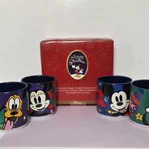 May include: A set of four ceramic mugs featuring Disney characters Pluto, Minnie Mouse, Mickey Mouse, and Goofy in holiday-themed designs, displayed in front of a red box labeled An Enchanted Christmas with a graphic of Mickey Mouse.
