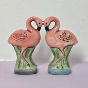 Op de afbeelding: Een paar keramische zout- en peperstrooiers in de vorm van flamingo's. Elke flamingo is roze met een zwarte snavel en staat op een groene basis met een blauwe onderkant. De vogels staan tegenover elkaar met hun nekken in een hartvorm.