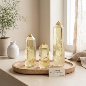 Könnte beinhalten: Drei polierte Citrin-Kristallspitzen unterschiedlicher Höhe auf einem ovalen Holztablett. Eine kleine rechteckige Karte davor trägt die Aufschrift Citrine Bring Joy & Abundance -SoulStrata. Die Kristalle sind durchscheinend gelb mit inneren Einschlüssen.
