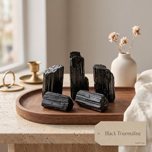 Puede incluir: Una colección de cinco cristales de turmalina negra en bruto con estrías verticales y bordes irregulares, dispuestos sobre una bandeja de madera lisa. Una pequeña etiqueta de papel muestra el texto Black Tourmaline y la marca SoulStrata.