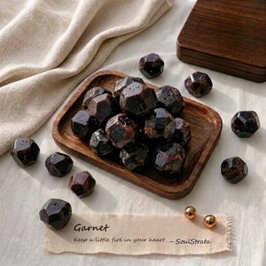 May include: A collection of dark red, faceted garnet stones piled on a small rectangular wooden tray, with several loose stones scattered nearby. A rectangular paper tag features the text "Garnet" and "Keep a little fire in your heart. - SoulStrata."