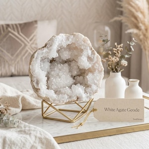Könnte beinhalten: Eine weiße Achat-Geode mit funkelnden kristallinen Innenformationen, die auf einem geometrischen goldfarbenen Metallständer ruht. Ein rechteckiges beiges Etikett mit goldener Schnur zeigt den Text White Agate Geode und den Markennamen SoulStrata.