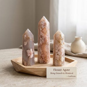 Può includere: Tre torri di cristallo in agata fiore lucidata di altezze variabili poggiano su un vassoio esagonale in legno. Le pietre presentano tonalità traslucide rosa, pesca e grigie con inclusioni bianche simili a fiori. Un cartellino riporta: Flower Agate Bring Growth & Renewal —SoulStrata.