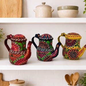 May include: Three metal teapots featuring hand-painted floral and geometric patterns in vibrant colors. The left teapot is maroon, the center is dark blue, and the right is yellow. Each teapot has a matching lid, a curved handle, and a spout.