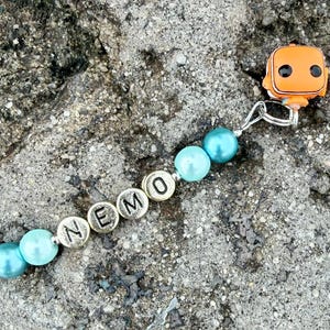May include: A beaded keychain featuring a small orange clownfish character charm attached to a silver metal ring. The design includes four light blue round beads and four silver-toned circular beads with the black embossed letters N-E-M-O.