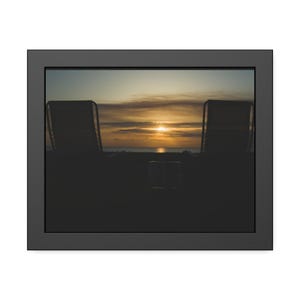 May include: A framed wall art print featuring a silhouette of two lounge chairs and a small table positioned against a sunset over the ocean. The scene is captured in dark tones with a bright golden sun glowing on the horizon beneath clouds.