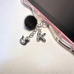 May include: A clear, glittery phone case features a dangling charm accessory attached to a silver metal ring. The charm includes a fluffy black pom-pom, a small silver letter K, and a miniature black and white character head with pink accents.
