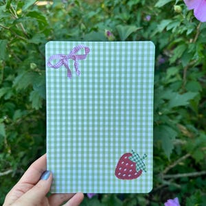 Puede incluir: Un bloc de notas rectangular con un patrón de cuadros vichy en verde claro y blanco. La esquina superior izquierda muestra un pequeño lazo vichy rosa, mientras que la esquina inferior derecha presenta una ilustración de fresa roja con una hoja vichy verde.