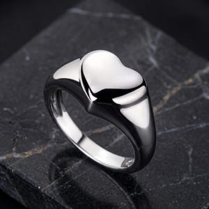 May include: A polished silver-tone ring featuring a smooth, raised heart-shaped centerpiece. The band is thick and rounded, tapering slightly toward the bottom, with a high-shine reflective finish that catches the light across its curved surface.