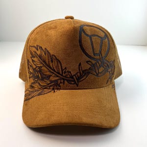 May include: A tan suede trucker hat featuring a dark brown ink illustration of a feather intertwined with barbed wire and a circular bull skull outline on the front panel. The cap has a structured front and a mesh back.