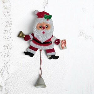 May include: A Santa Claus ornament featuring a red and white suit, a green holly accent on the hat, and a gold bell in one hand. The figure holds a small white sign with the red text Merry Christmas and has a dangling silver bell attached below.