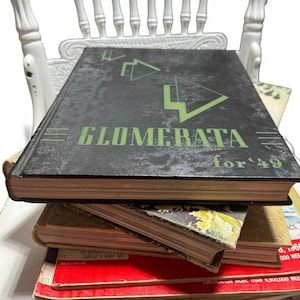 May include: A stack of vintage books and magazines on a white chair. The top book is black with green text that reads "GLOMERATA for '49". Other books and magazines are visible, with red and tan covers.