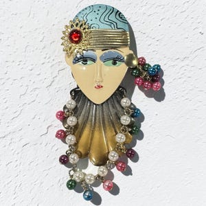 May include: A decorative brooch featuring a stylized face with a blue patterned headpiece, a gold-tone flower with a red center, and a scalloped collar. The piece is adorned with dangling multi-colored faux pearl beads in white, pink, green, and blue.