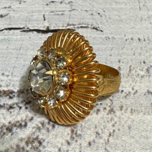 May include: A gold-toned ring featuring a large central clear faceted stone surrounded by a cluster of smaller clear stones. The setting is composed of numerous thin, radiating gold-toned wire coils that create a textured, circular frame.