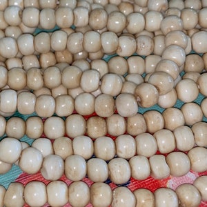 May include: Close-up of strands of off-white, oval-shaped beads. The beads are strung together, creating multiple rows. The beads have a smooth, glossy finish and are uniform in size. The background is a colorful floral pattern.