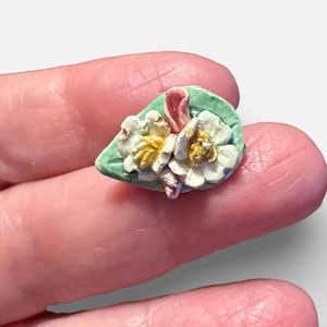 May include: A miniature sculpture of white flowers with yellow centers and pink accents, set on a green leaf-shaped base. The delicate floral arrangement is a small, detailed piece of art.