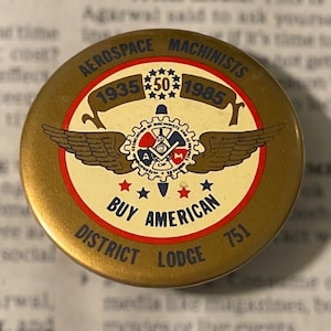 May include: A gold-colored button with a red, white, and blue design. It says "Aerospace Machinists 1935 50 1985" and "Buy American" with a winged gear in the center. The bottom says "District Lodge 751".