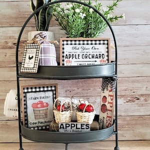 May include: A two-tiered, dark metal serving tray with farmhouse-style decor. The top shelf holds a small potted plant, a sign that reads "Pick Your Own Apple Orchard," and a gingham-wrapped item. The bottom shelf features signs, baskets of apples, and the text "Apples 25¢ a Bushel."