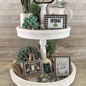 May include: A two-tier white tiered tray decorated for St. Patrick's Day. The top tier features a green shamrock, a white pitcher, and a sign that says "Lucky". The bottom tier features a small potted plant, a sign that says "Hainlen's Irish Pub", a sign that says "Kiss Me I'm Irish", and a sign that says "March Pot of Gold".