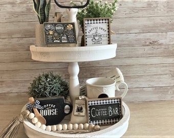 May include: Two-tiered white wooden tray with a variety of coffee-themed decorations. The top tier features a potted plant, a sign that says "Coffee Shop Lounge", and a sign that says "Coffee House". The bottom tier features a white mug, a black mug with the words "Coffee House", a wooden spoon, a wooden bead garland, a sign that says "Coffee Bar", and a small tag with a coffee bean design.