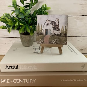 May include: A small, square painting of a white barn with a silo, set on a miniature wooden easel. The artwork depicts a rural scene with a path leading to the barn, surrounded by trees and a fence. The painting is displayed on a stack of books.