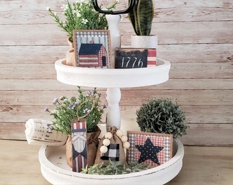 May include: A two-tier white wooden tiered tray decorated with patriotic themed decor. The top tier features a small potted plant, a burlap bag, and a wooden sign with the number 1776. The bottom tier features a small potted plant, a burlap bag with wooden beads, a wooden sign with a star, and a wooden sign with a figure wearing a red, white and blue outfit.