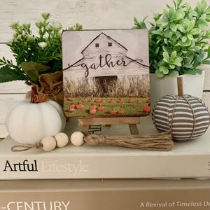 May include: A square wooden sign featuring a white barn illustration with the word gather in black script, set on a small wooden easel. The scene includes a field of orange pumpkins. A jute tassel with two wooden beads sits in front of the sign.