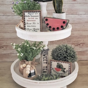May include: A white tiered tray with two levels decorated with summer-themed items. The top tier features a watermelon slice, a potted succulent, and a sign that reads "Hello Summer Ice Cold 50c Watermelon Sweet Tea 25c Bike Rides Picnic Baskets". The bottom tier features a potted succulent, a burlap bag with white beads, a sign that reads "Sweet Tea", and a sign that reads "Watermelons".