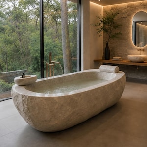 Puede incluir: Una bañera ovalada independiente tallada en piedra natural de color beige claro con un exterior rugoso y texturizado y un interior liso. La bañera cuenta con un reposacabezas de piedra a juego y una pequeña bandeja integrada con una botella.