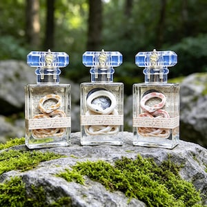 May include: Three rectangular glass perfume bottles with blue faceted caps and gold snake accents. Each bottle contains a coiled snake figure and a paper label with cursive text wrapped around the center, displaying orange, white, and pink snakes.