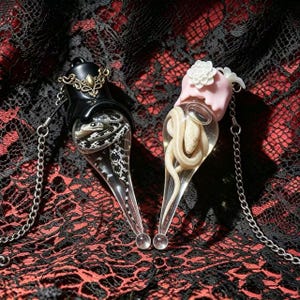 May include: Two teardrop-shaped glass pendants on silver chains. One pendant features a black cap with gold filigree and a black and white snake inside. The other features a pink cap with a white flower and a cream-coloured snake inside.