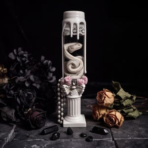 May include: A clear glass cylinder containing a coiled white snake figure, topped with white dripping wax and lace. The cylinder rests on a white classical column base adorned with pink floral accents, a central gem, and a dangling string of white pearls.