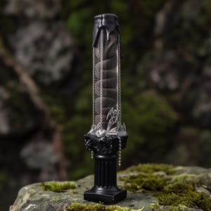 May include: A cylindrical glass container filled with a dark coiled snake-like object, topped with black wax drips. The base is a black fluted column pedestal adorned with silver chains and small metal charms including a heart and a scythe.