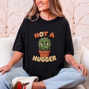 Pode incluir: Uma t-shirt preta de manga curta com um gráfico de um cacto verde num vaso castanho, com uma expressão rabugenta e braços cruzados. O texto acima do cacto diz NOT A e o texto abaixo do cacto diz HUGGER em letras de estilo negrito.