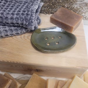 May include: A dark olive green ceramic soap dish with five drainage holes, a rectangular brown soap bar, a folded grey waffle-textured cloth, and several square soap bars in shades of tan, beige, and cream arranged on a light wood surface.