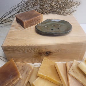 May include: A collection of rectangular soap bars in various shades of brown, tan, and yellow, accompanied by a small, dark green, oval-shaped ceramic soap dish with drainage holes, all arranged on a light-colored wooden block surface.