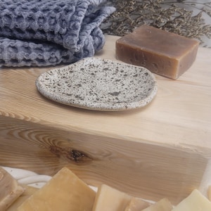 May include: A speckled ceramic soap dish with an irregular oval shape, featuring a cream-colored base with dark brown flecks. The dish is positioned near a grey waffle-weave cloth and a rectangular bar of brown soap, with additional soap bars below.