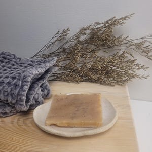 May include: A rectangular bar of beige soap with visible flecks of botanical material rests on a small, round, off-white ceramic dish. Beside the dish is a folded grey textured fabric and a sprig of dried brown flowers.