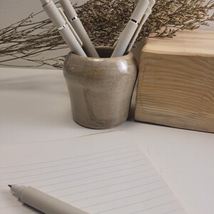 May include: A light beige ceramic pen holder containing four matching light beige pens with silver accents. A single light beige pen rests on a lined notebook in the foreground, accompanied by a wooden block and dried decorative foliage.